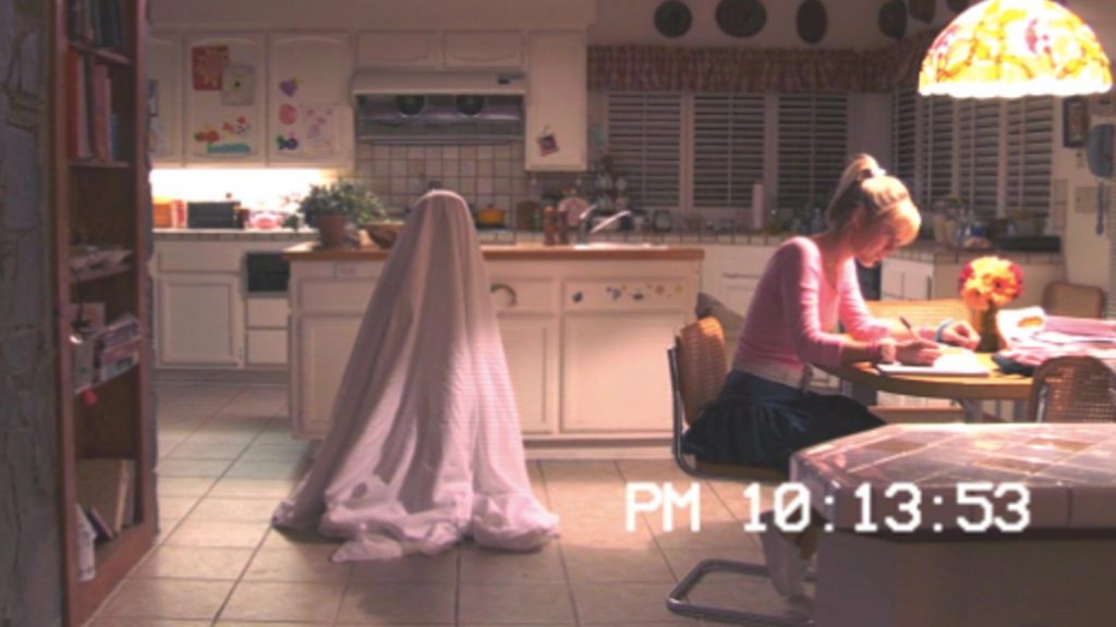 Un adolescente se sienta en una mesa de cocina haciendo la tarea mientras un niño cubierto con una sábana está detrás de ella